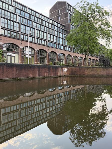 This photo captures a modern brick building with numerous windows and arched ground-floor entrances, reflected perfectly in the calm waters of a canal at Plantage Kerklaan, Plantage, Amsterdam